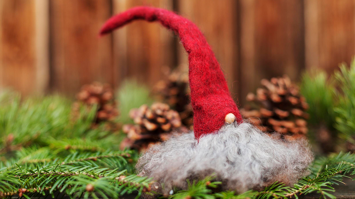 Jultomte av ull med tovad tomteluva, som sitter på granris och det är flera kottar i bakgrunden. 