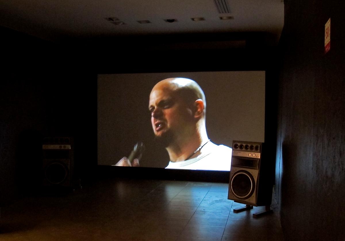 I ett mörkt rum står en projektorskärm som visar en skalig man i hip-hopar stil som håller i en mick. På sidan av skärmen står två högtalare. 