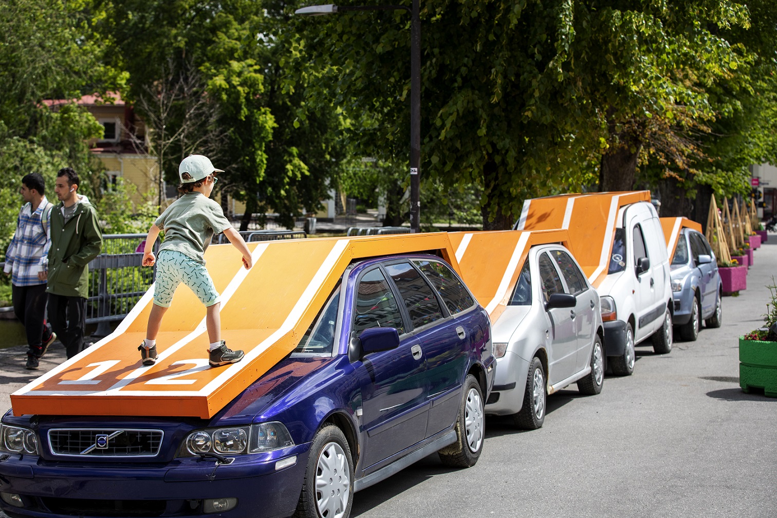 Outside there is four cars of different sizes and models are lined up on a street. Above the cars, the artist has built an orange running track with two tracks that follow the roof and shapes of the cars. The artwork opens up to the idea of a running race over the cars and in the picture a child has climbed onto the running track and is ready.
